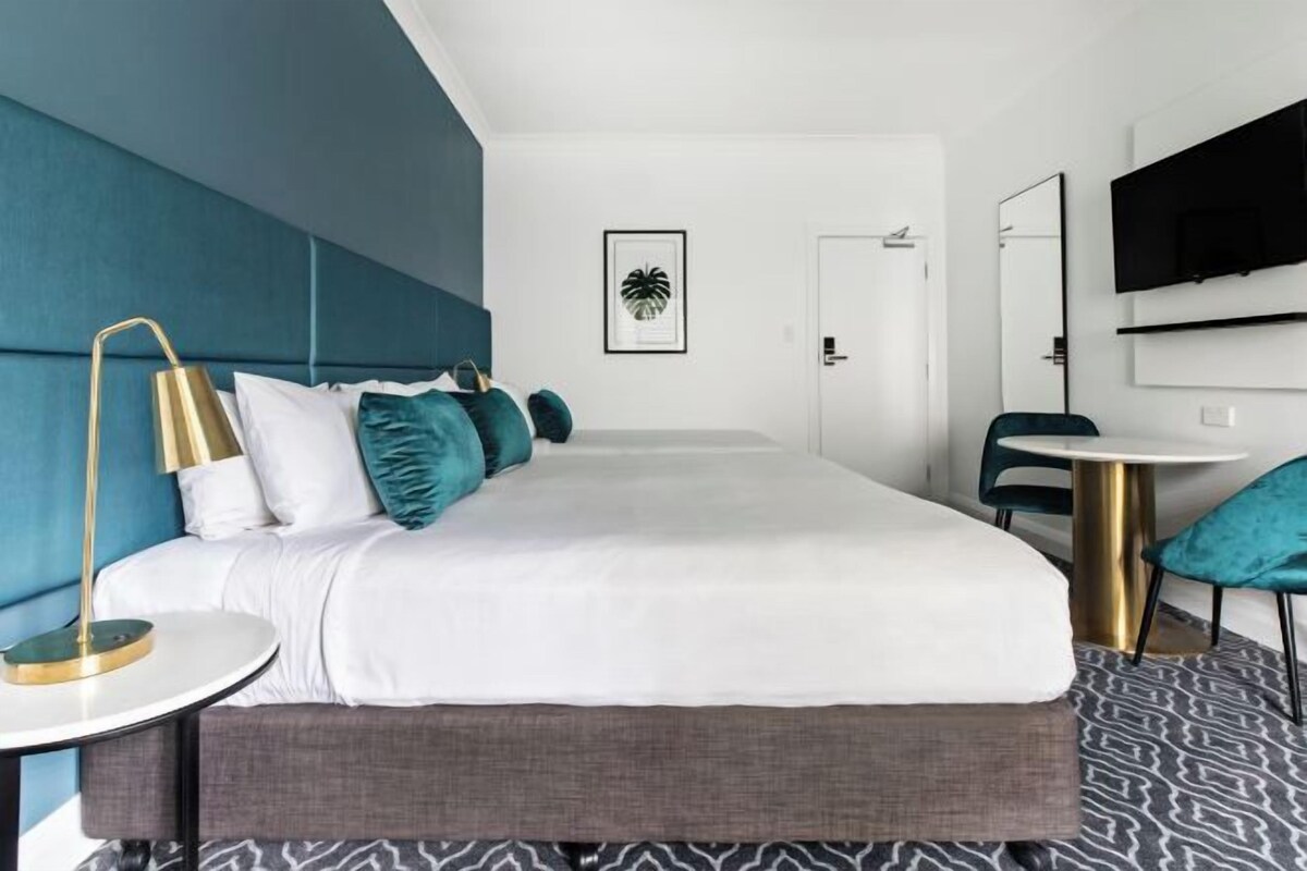 A modern hotel room with a large bed, a round table, a TV mounted on the wall, and a door leading to another room.