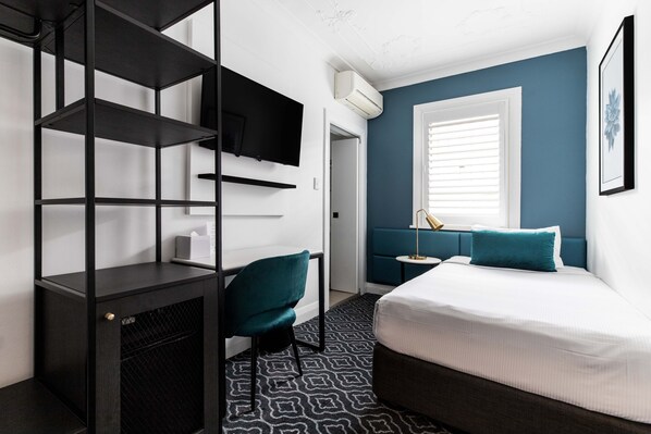 A modern hotel room with a bed, a desk with a chair, a TV mounted on the wall, and a window with blinds.