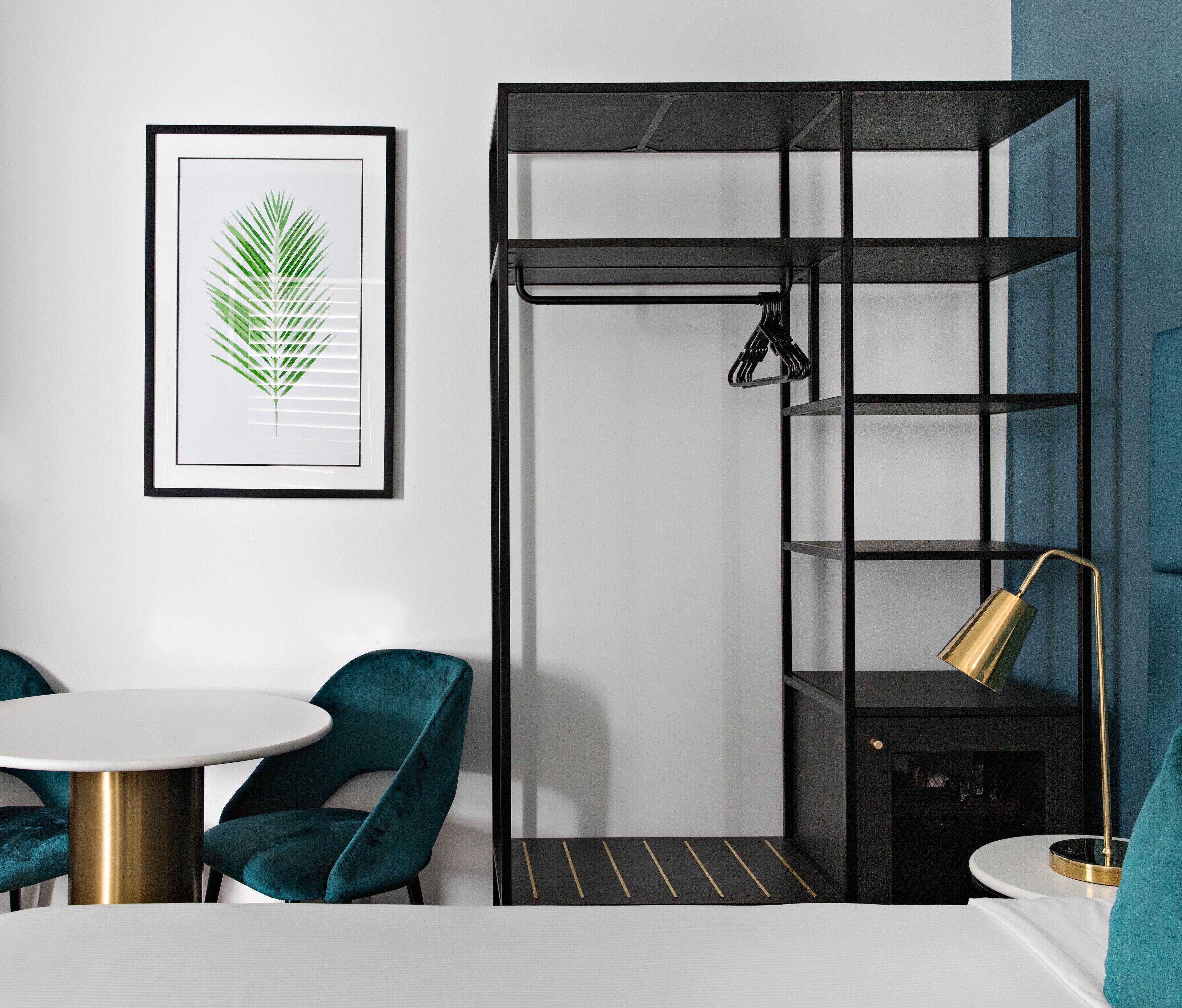 A compact room with a bed, a small round table, teal chairs, a framed leaf artwork, a black shelving unit with a hanging lamp, and a gold lamp on a bedside table.