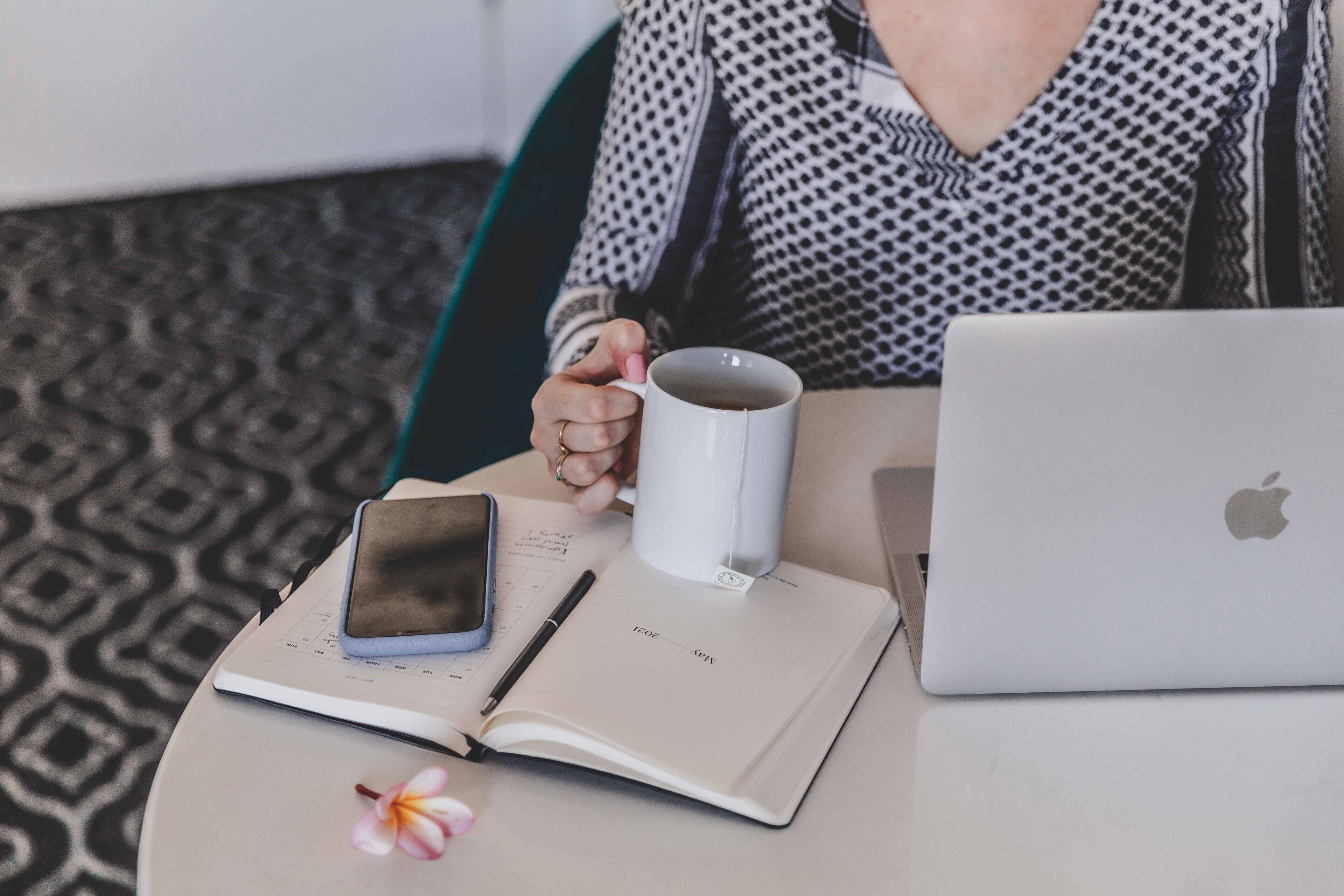 Une personne assise à une table, devant un ordinateur portable, avec un carnet, un téléphone intelligent, une tasse et un stylo.