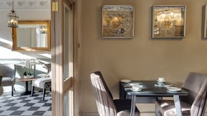 Un comedor con piso a cuadros, una mesa dispuesta para dos personas y dos sillas. Se observan obras de arte enmarcadas en la pared y un candelabro sobre la mesa.