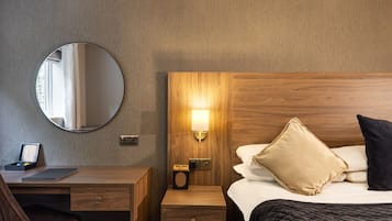 Une chambre d’hôtel avec une tête de lit en bois, une table de chevet avec une lampe, un miroir rond, un bureau avec un ordinateur portable et un lit avec des oreillers.