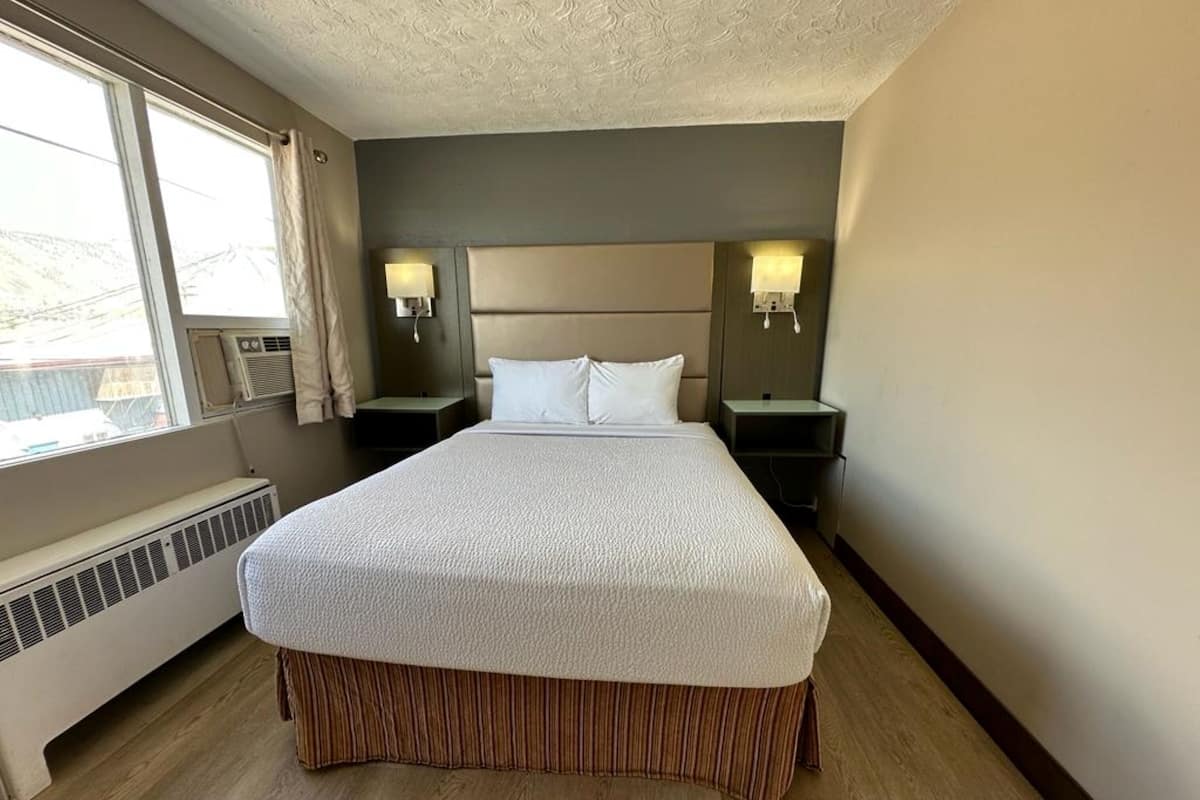 A hotel room with a large bed, two bedside tables, a window with curtains, and a built-in heater.