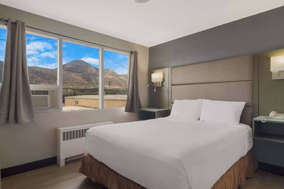A hotel room with a large bed, a nightstand, a lamp, a window with curtains, and a view of mountains.