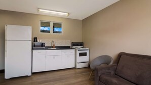 A compact kitchen with white cabinets, a microwave, a coffee maker, and a stove. There's a window above the sink and a gray chair in the living area.