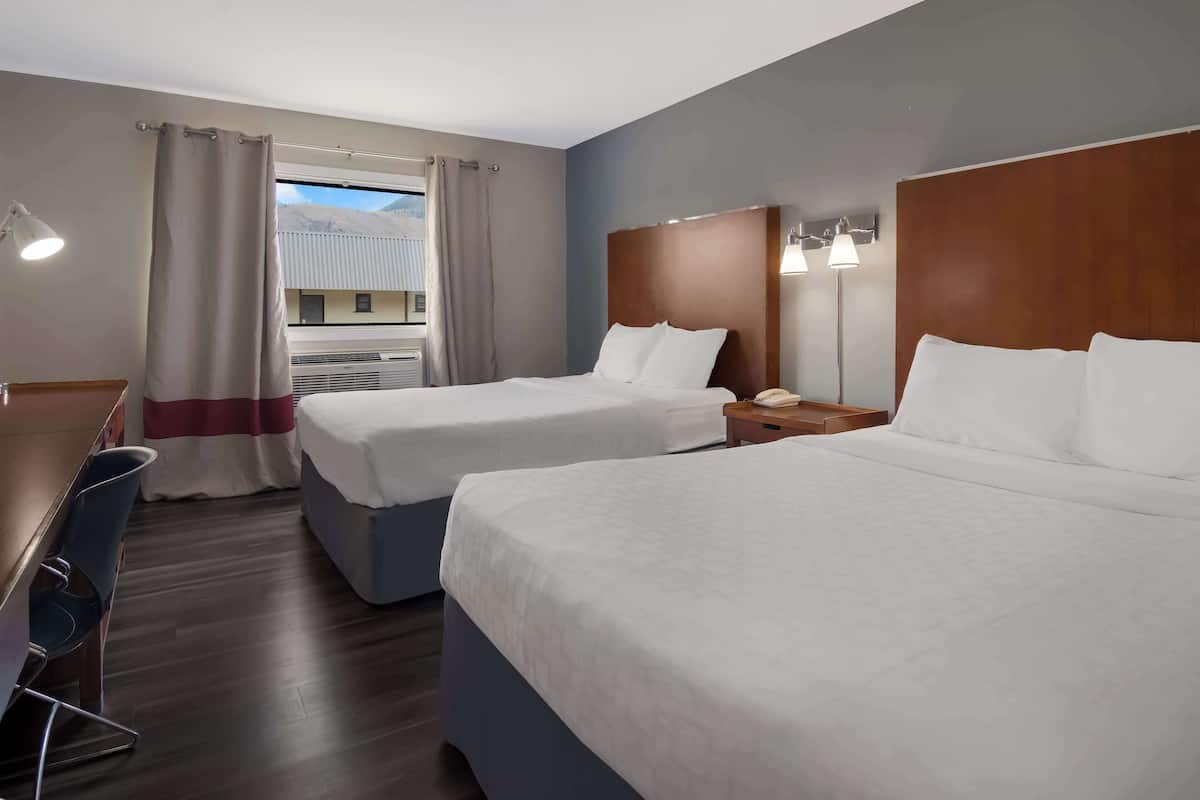 A hotel room with two beds, a desk, and a chair. There is a window with curtains, a nightstand with a lamp, and a view of mountains.