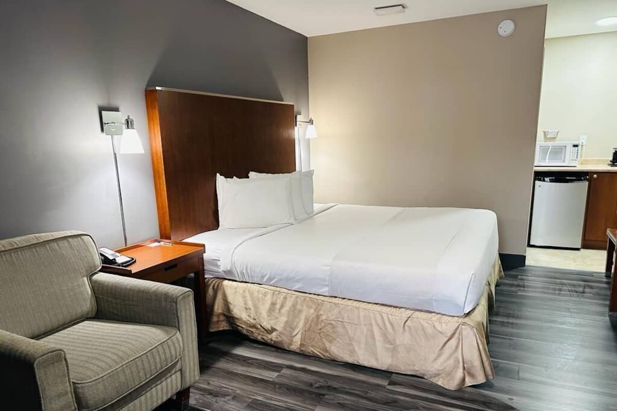A hotel room with a bed, a chair, a desk with a lamp, and a kitchenette.