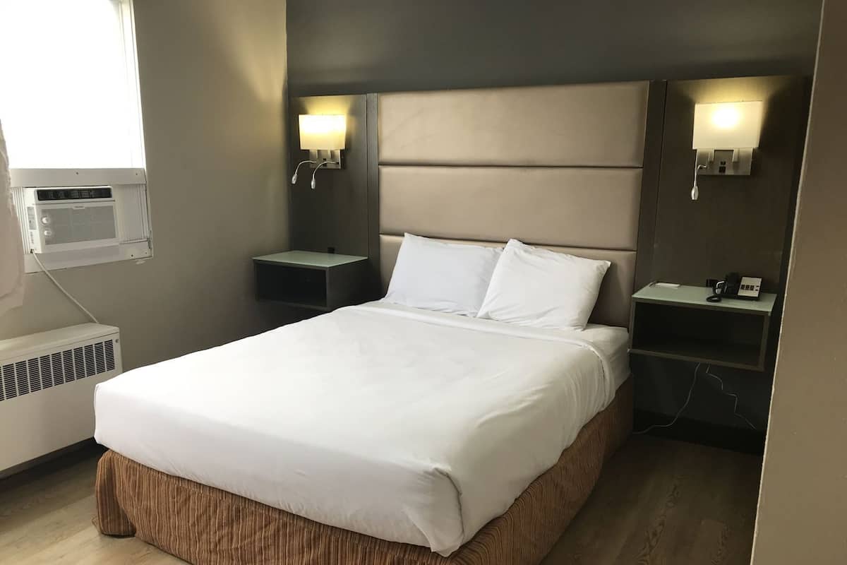 A neatly made bed in a hotel room with a headboard, two bedside tables, a wall-mounted air conditioner, and a small desk with a telephone.
