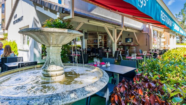 An outdoor dining area with a stone fountain, tables, and chairs, and a restaurant named 'Semys' in the background. - Logis Hôtel Noemys Brive - Restaurant Le Teinchurier (Brive-la-Gaillarde)