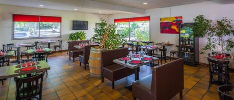 Un restaurante con varias mesas y cabinas, ventanas amplias con persianas rojas y plantas de interior.