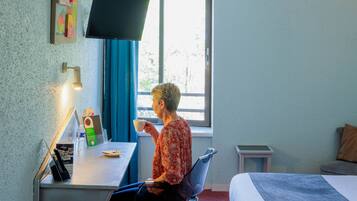 A hotel room with a desk, chair, bed, TV, and window with curtains.