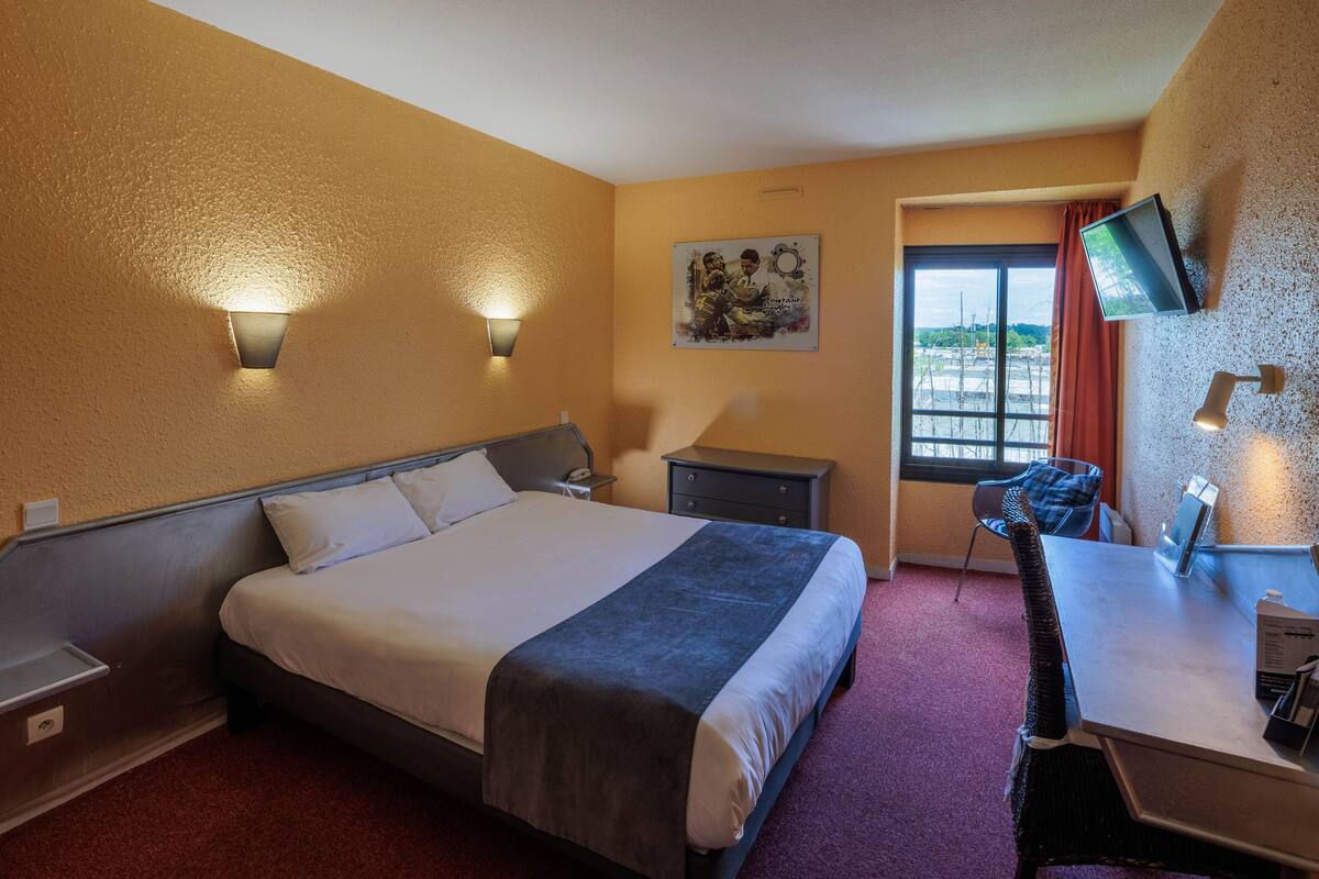 Habitación de hotel con cama, mesitas de noche, escritorio, silla, televisor montado en la pared y ventana con cortinas.