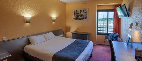 Une chambre d’hôtel comprenant un lit, des tables de chevet, un bureau, une chaise, un téléviseur fixé au mur et une fenêtre avec des rideaux.