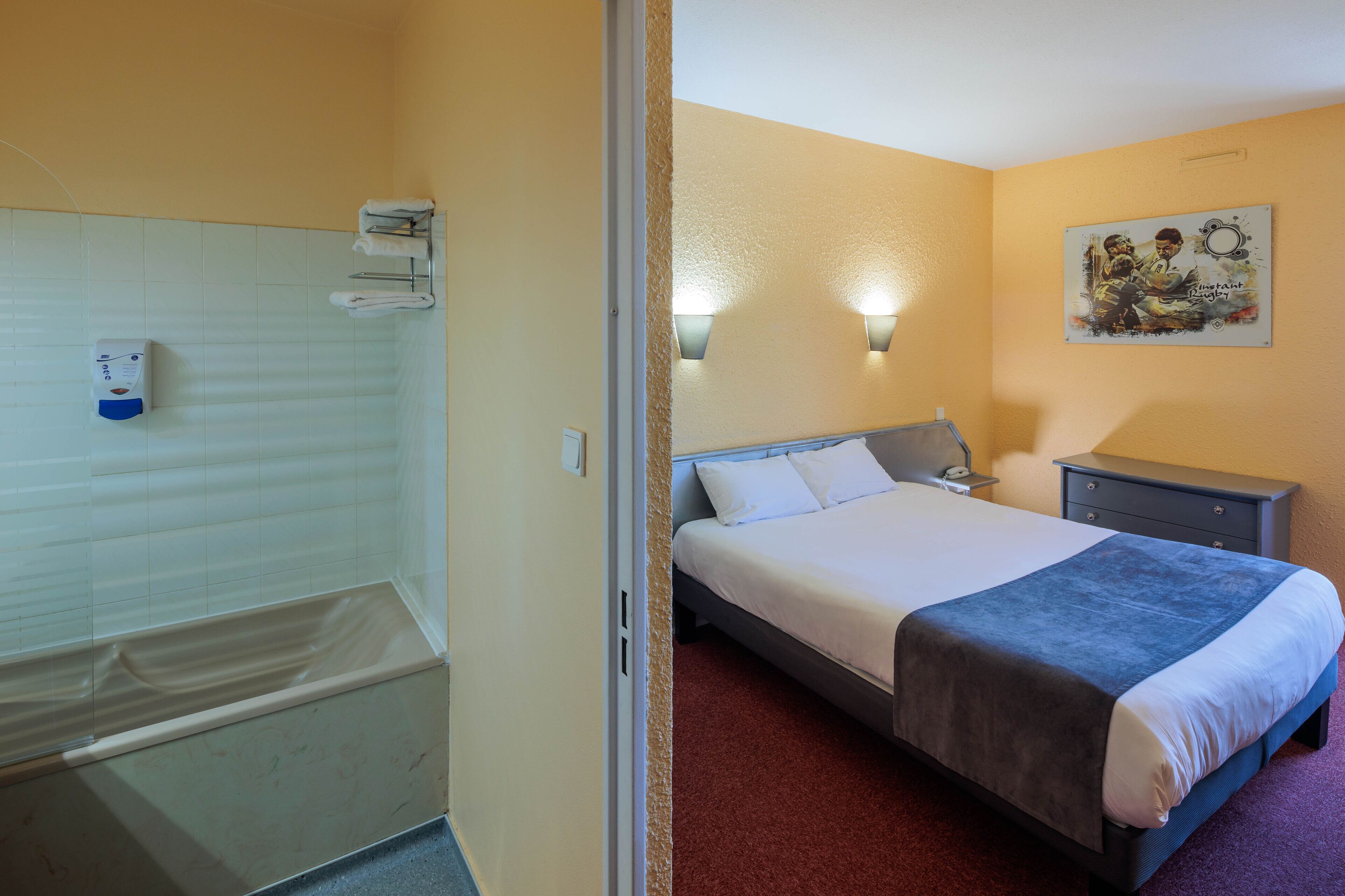 A hotel room with a bathtub, a bed with a blue blanket, a nightstand, and a wall-mounted light fixture.