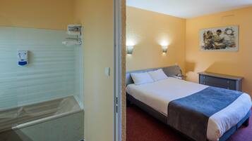 A hotel room with a bathtub, a bed with a blue blanket, a nightstand, and a wall-mounted light fixture.