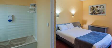 A hotel room with a bathtub, a bed with a blue blanket, a nightstand, and a wall-mounted light fixture.