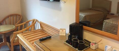 A hotel room with a bed, a desk with a coffee maker, a TV mounted on the wall, and a chair.