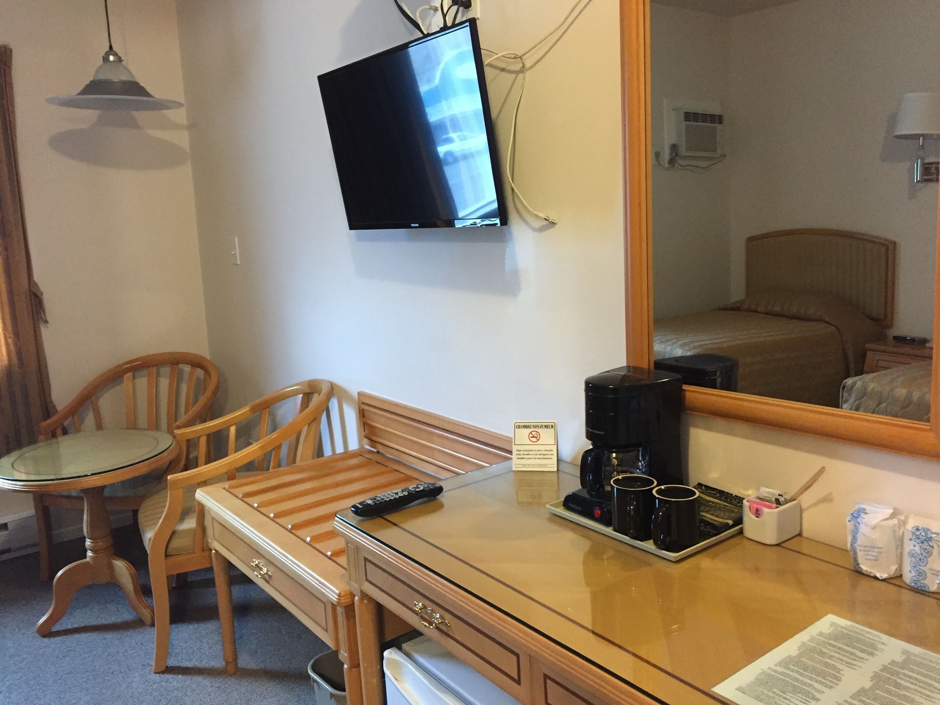 A hotel room with a wooden desk, a chair, a television mounted on the wall, a coffee maker, and a mirror reflecting the room.