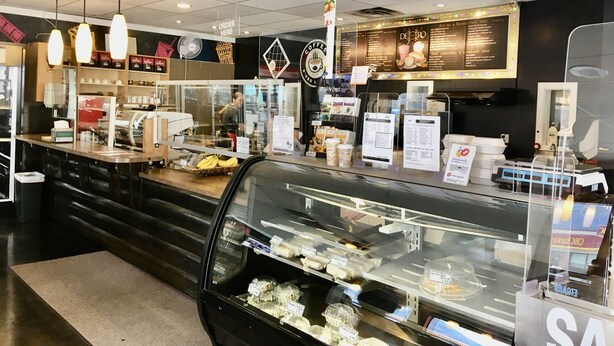 Interior de una cafetería con mostrador, cartel de menú y diversos productos alimenticios a la vista.