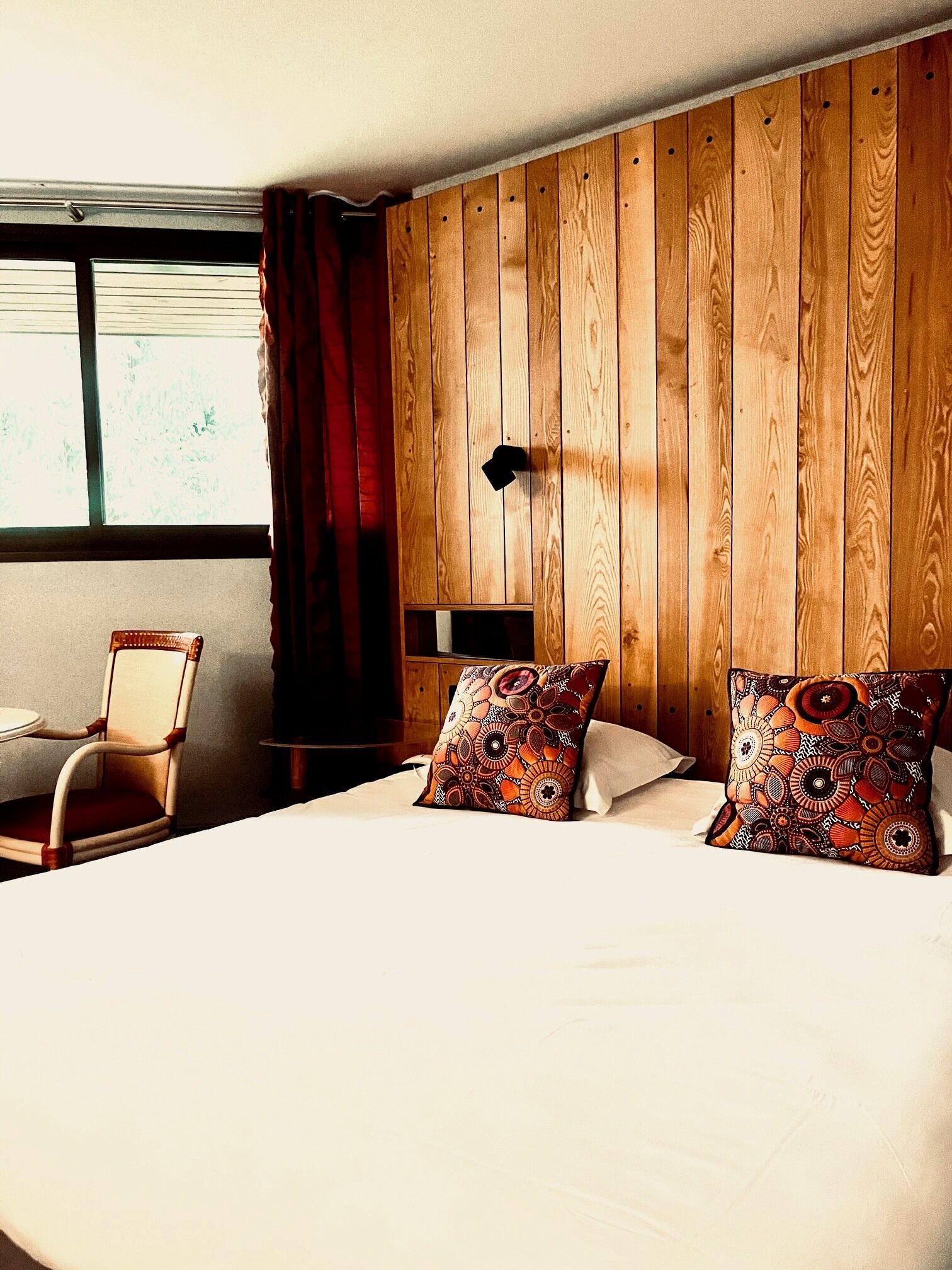 Habitación de hotel con pared de paneles de madera, cama con almohadones estampados, una silla y una ventana con cortinas.