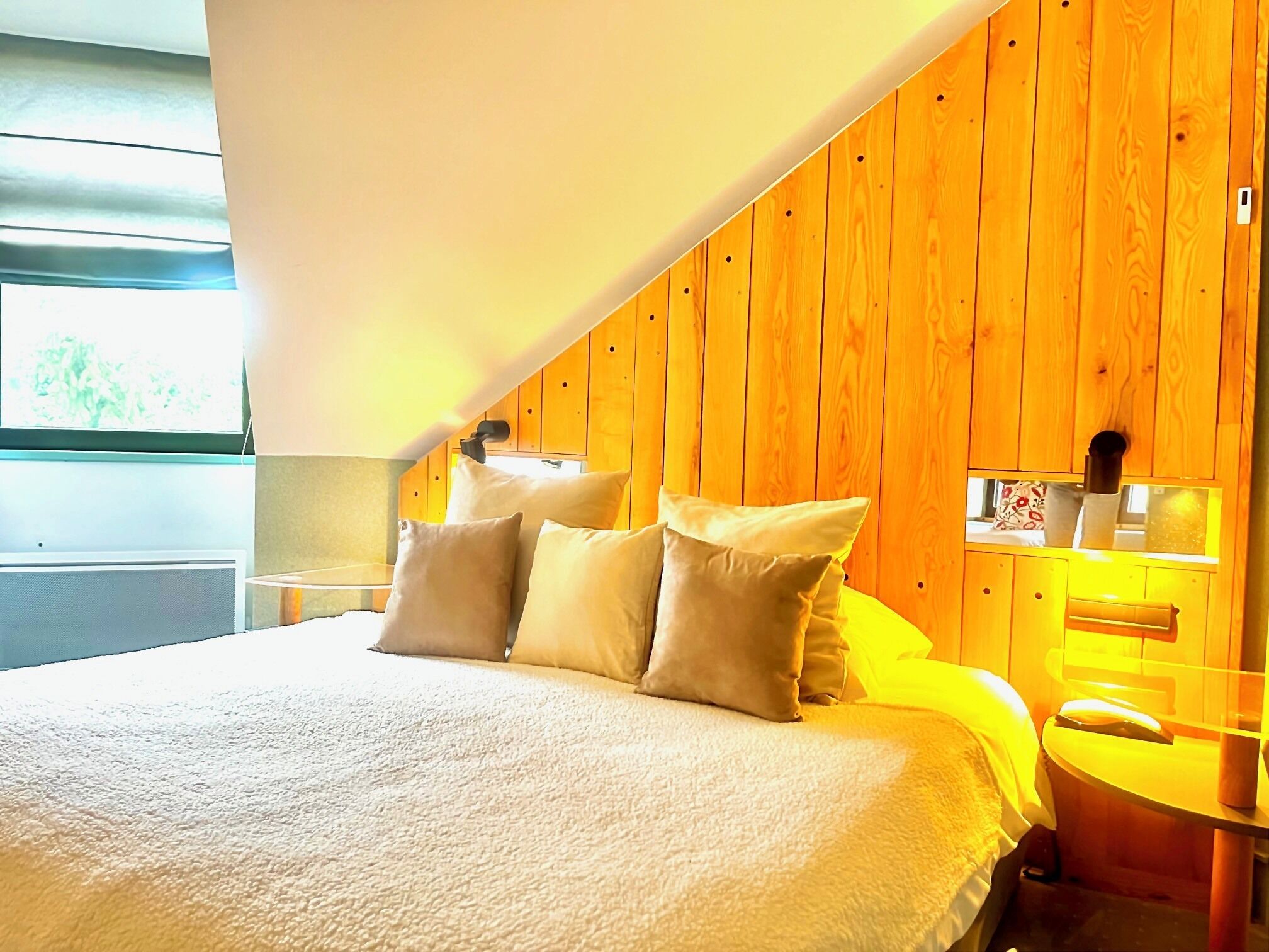 Un dormitorio con techo inclinado de madera, una cama con ropa de cama blanca y una mesita de noche con lámpara.