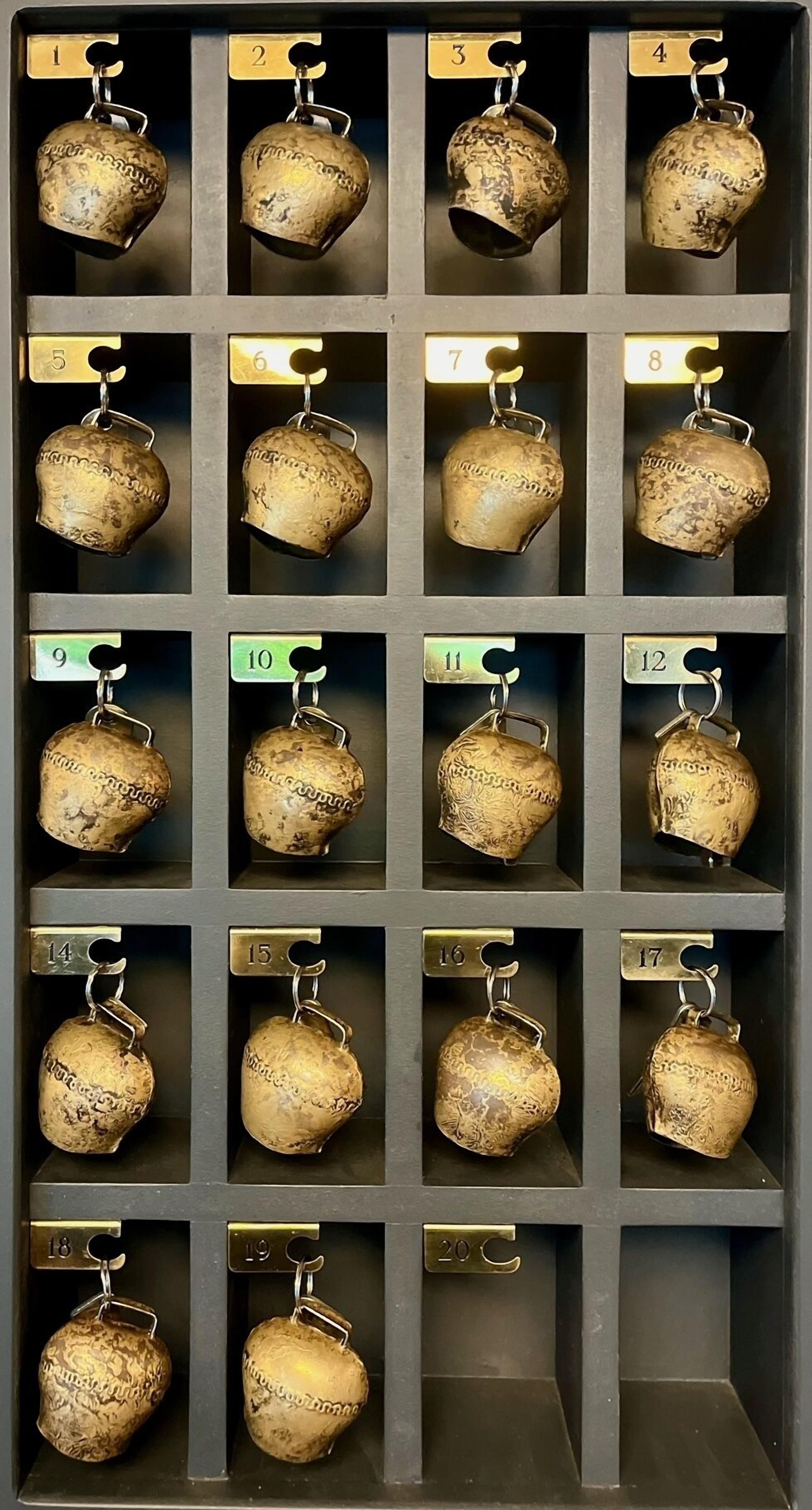 Une présentation de 20 objets métalliques en forme de cloche, numérotés et dotés d’une texture particulière, disposés en grille.