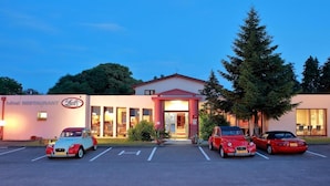A hotel with a red and white facade, a parking lot with three cars, and a large tree to the right. - Le Luth (Mirecourt)