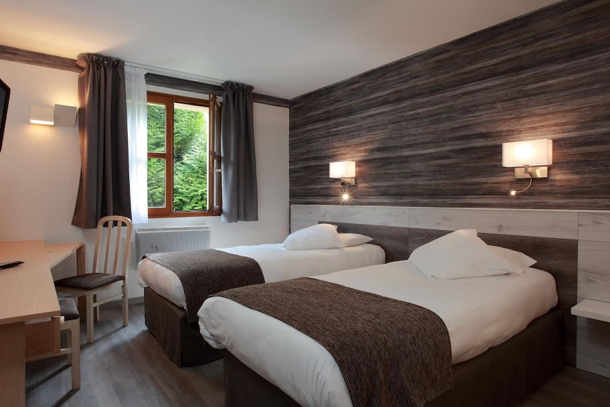 A hotel room with two beds, a desk, a chair, a TV, and a window with curtains.