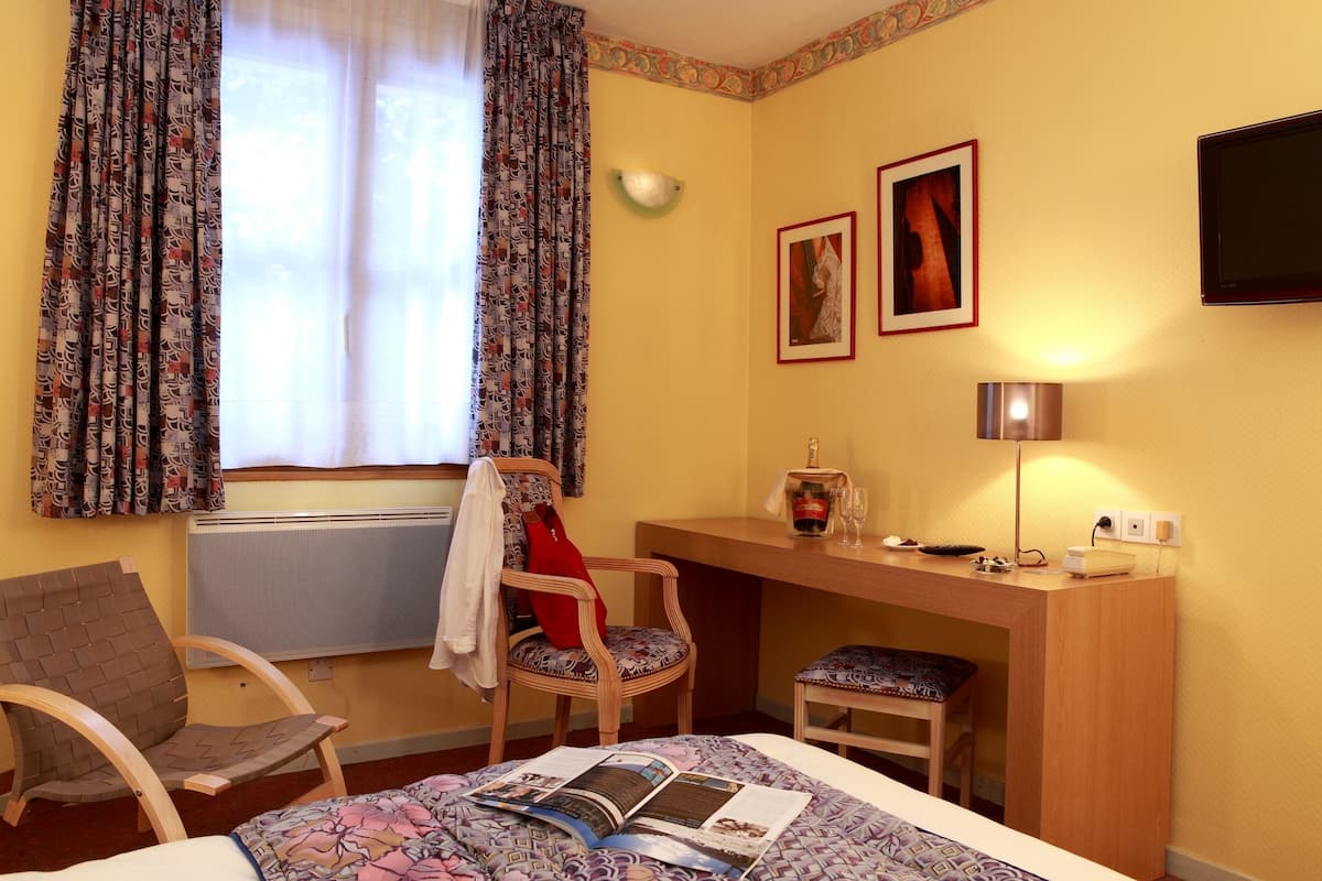 A hotel room with a bed, a desk, a chair, a window with curtains, a wall-mounted TV, and two framed pictures.