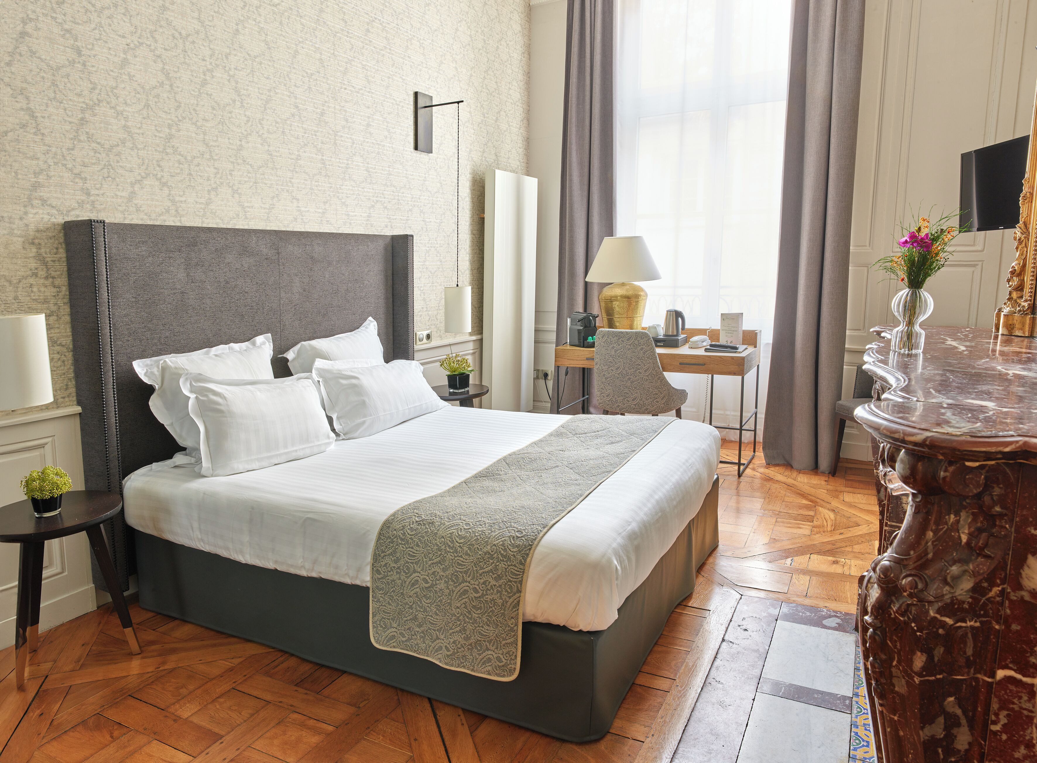 A hotel room with a large bed, a desk with a lamp, a TV, and a vase with flowers.