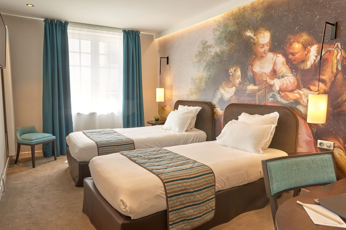 A hotel room with two beds, a desk, a chair, a lamp, and a large painting on the wall.