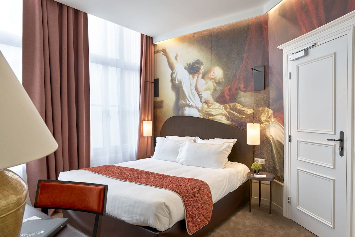 A hotel room with a large bed, a desk with a laptop, a nightstand with a lamp, and a door.