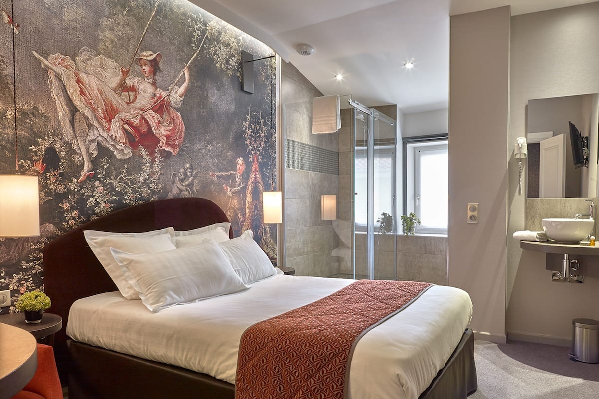 A modern hotel room with a large bed, a glass shower, a bathroom with a sink and mirror, and a mural on the wall.