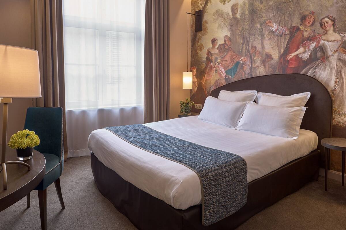 A hotel room with a large bed, a desk with a lamp, a chair, and a painting on the wall.