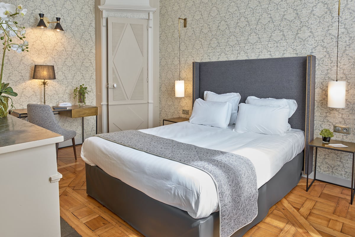 A modern bedroom with a large bed, bedside tables, a desk, and a chair. There are two wall-mounted lamps and a door with a decorative design.