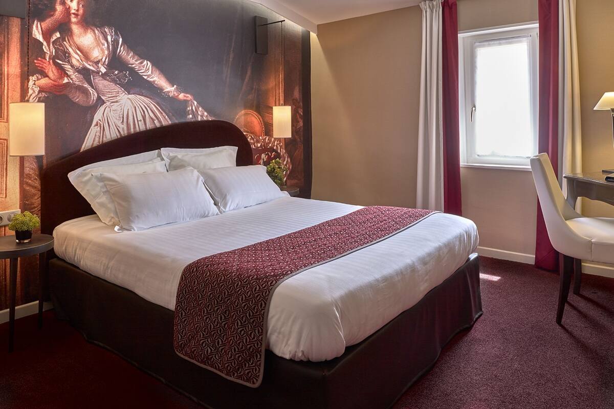 A hotel room with a large bed, a desk, a chair, a window with curtains, and a wall mural.