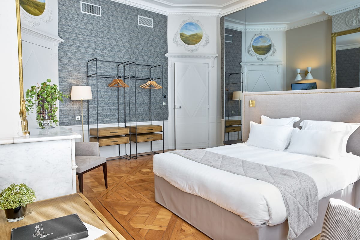 A modern hotel room with a large bed, a desk with a lamp, a mirror, and a wardrobe.