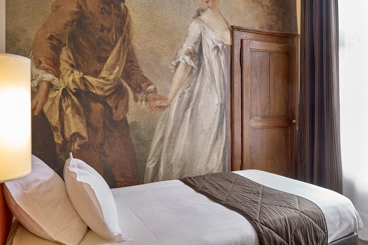 A hotel room with a bed, a wooden wardrobe, and a wall painting.