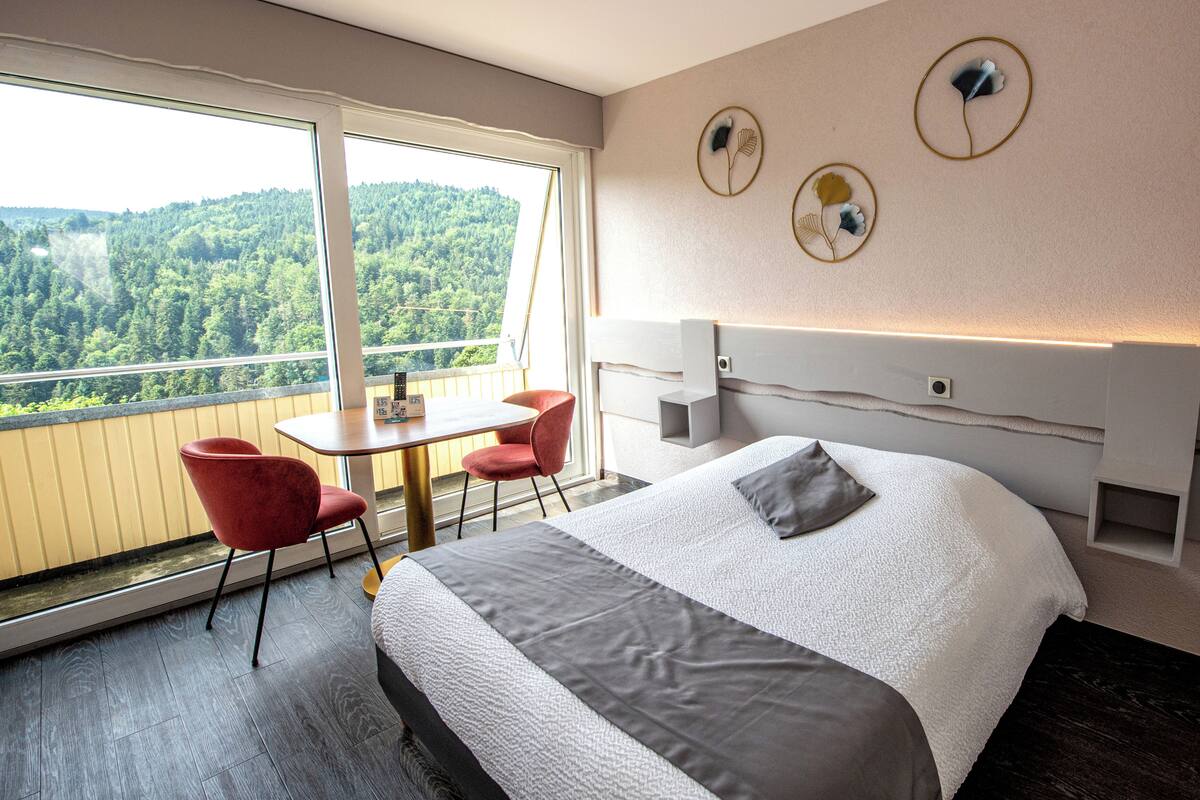 Habitación de hotel con una cama grande, una mesita con dos sillas, un balcón con vistas a una zona boscosa y decoración en las paredes.