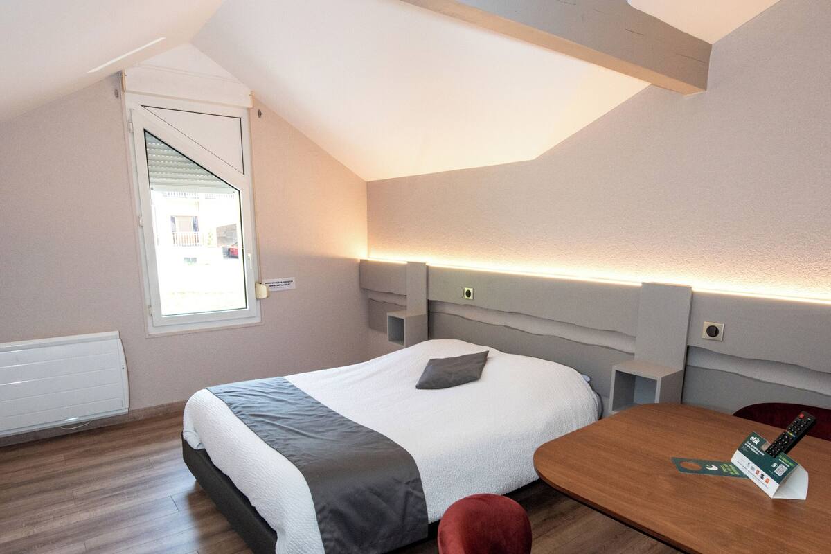 Una habitación de hotel moderna con una cama, una silla roja, una mesa de madera y un tragaluz.