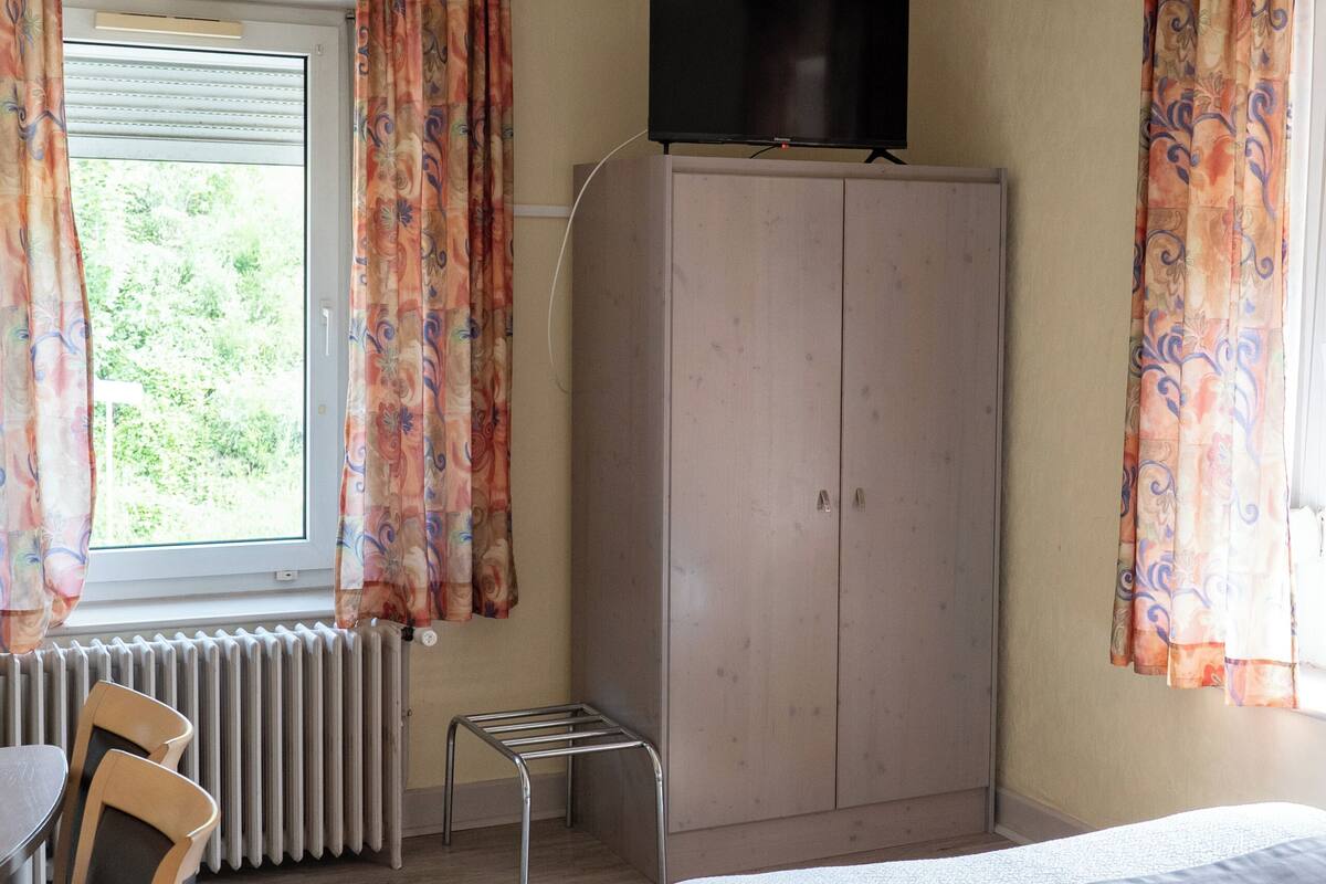 Un dormitorio con cama, televisor en un mueble, radiador, una silla y cortinas.