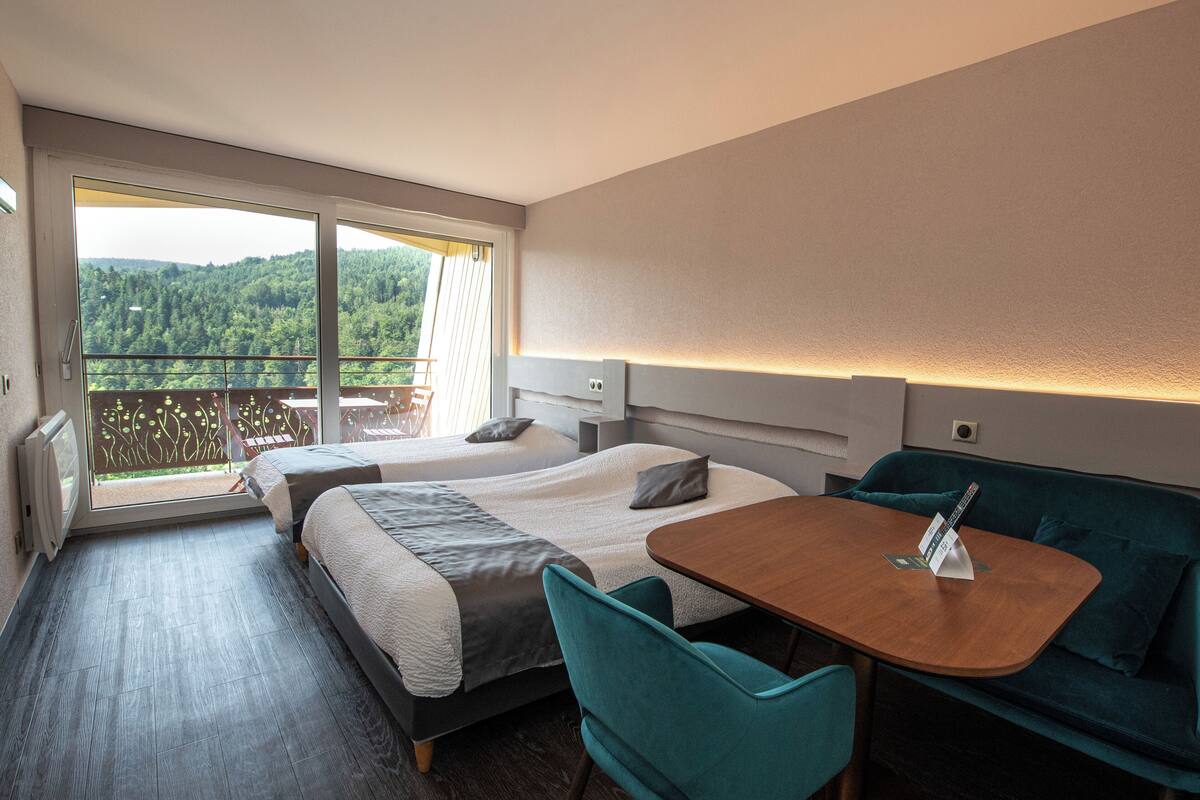 Habitación de hotel con dos camas, un balcón con vistas, una mesa de comedor y sillas color turquesa.