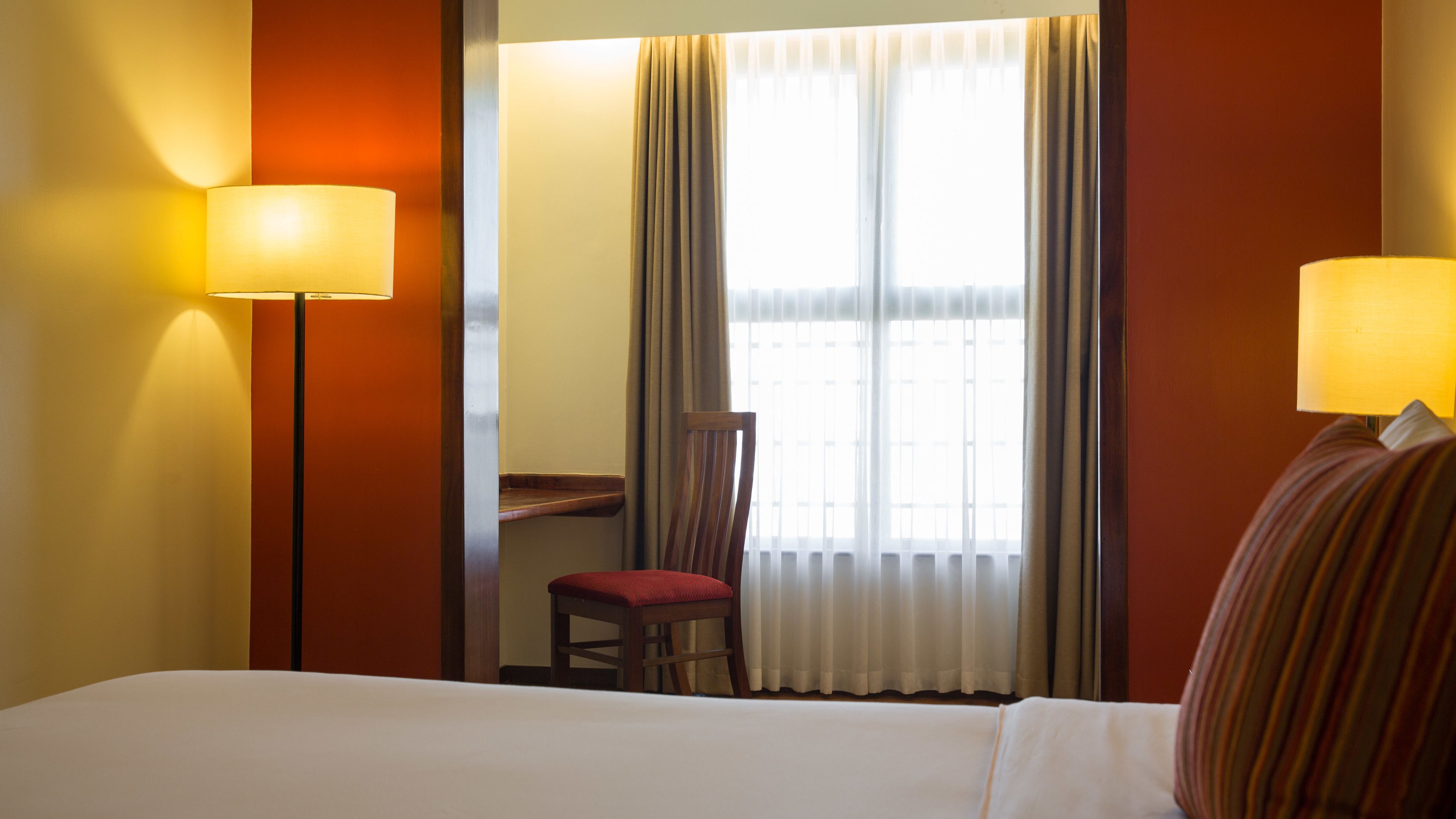 Ett hotellrum med en säng, två lampor, en stol och ett fönster med gardiner.
