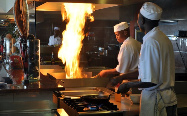 A kitchen with chefs preparing food, a large flame, and cooking equipment. - Ole Sereni (Nairobi)