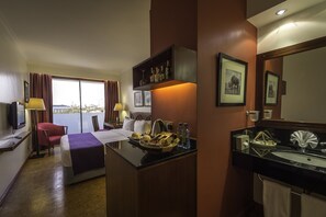A hotel room with a bed, a desk with a lamp, a bathroom with a sink and mirror, and a window with curtains. - Ole Sereni (Nairobi)