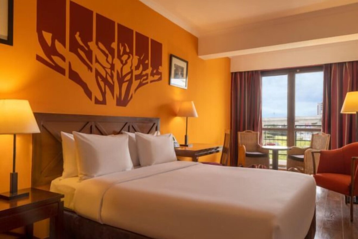 A hotel room with a large bed, two bedside lamps, a headboard, a window with curtains, a small table with chairs, and a view of the city.