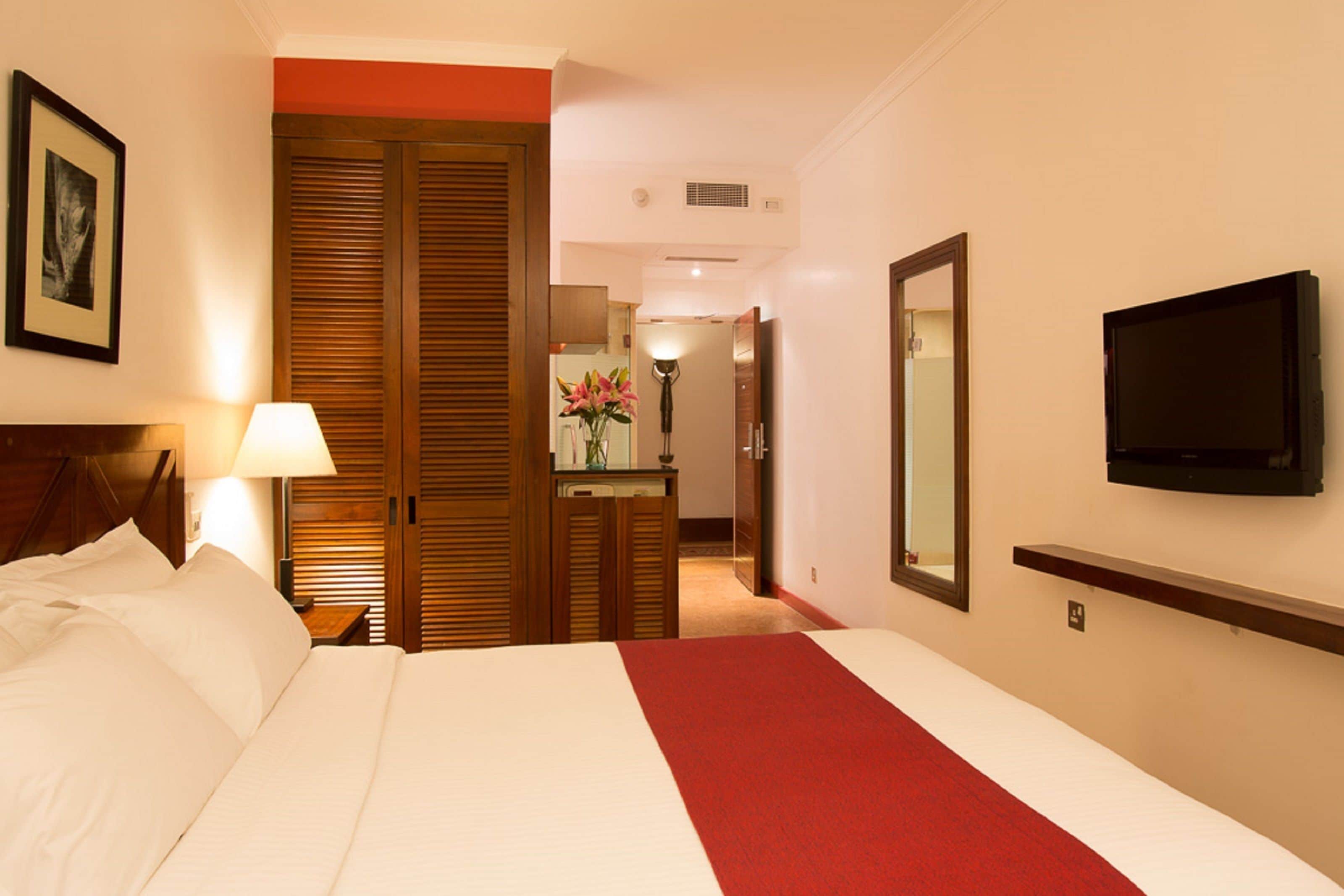 Une chambre d’hôtel avec un grand lit, un bureau, un miroir et un téléviseur fixé au mur.