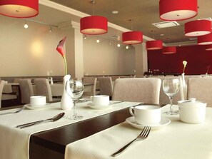 Un espace repas avec des tables dressées, des lampes suspendues rouges et un vase contenant une seule fleur.