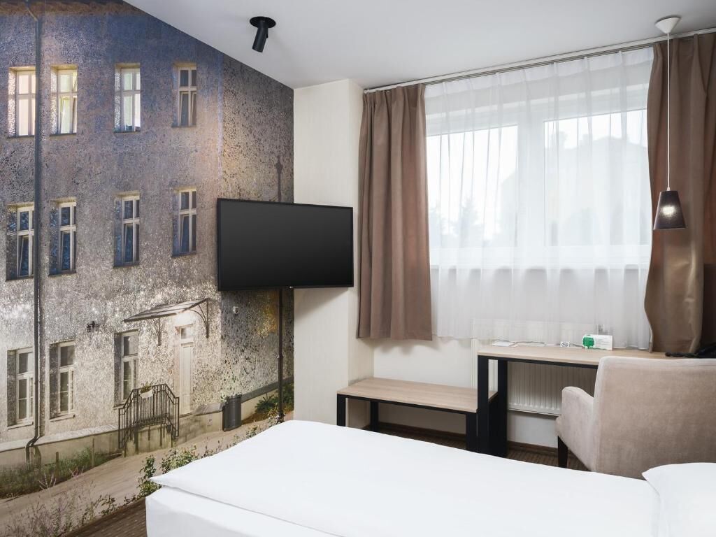 Une chambre d’hôtel comprenant un lit, un bureau, une chaise, un téléviseur fixé au mur et une grande fenêtre avec des rideaux.