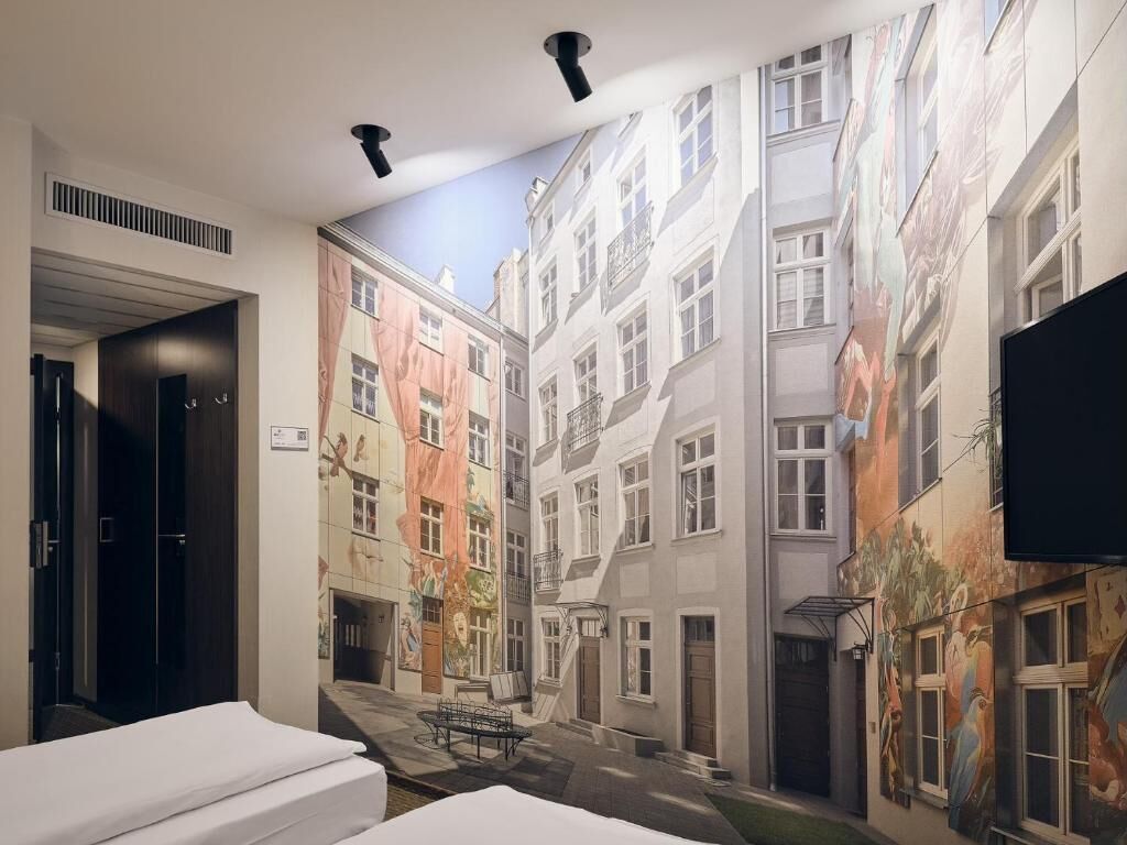 A hotel room with a mural of a European street, a bed, a desk, and a TV.
